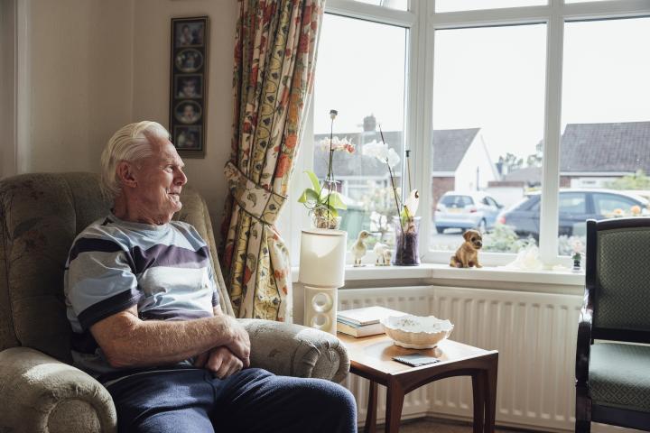 side view of a senior man sitting in a living room at his home looking out of his front window onto the street