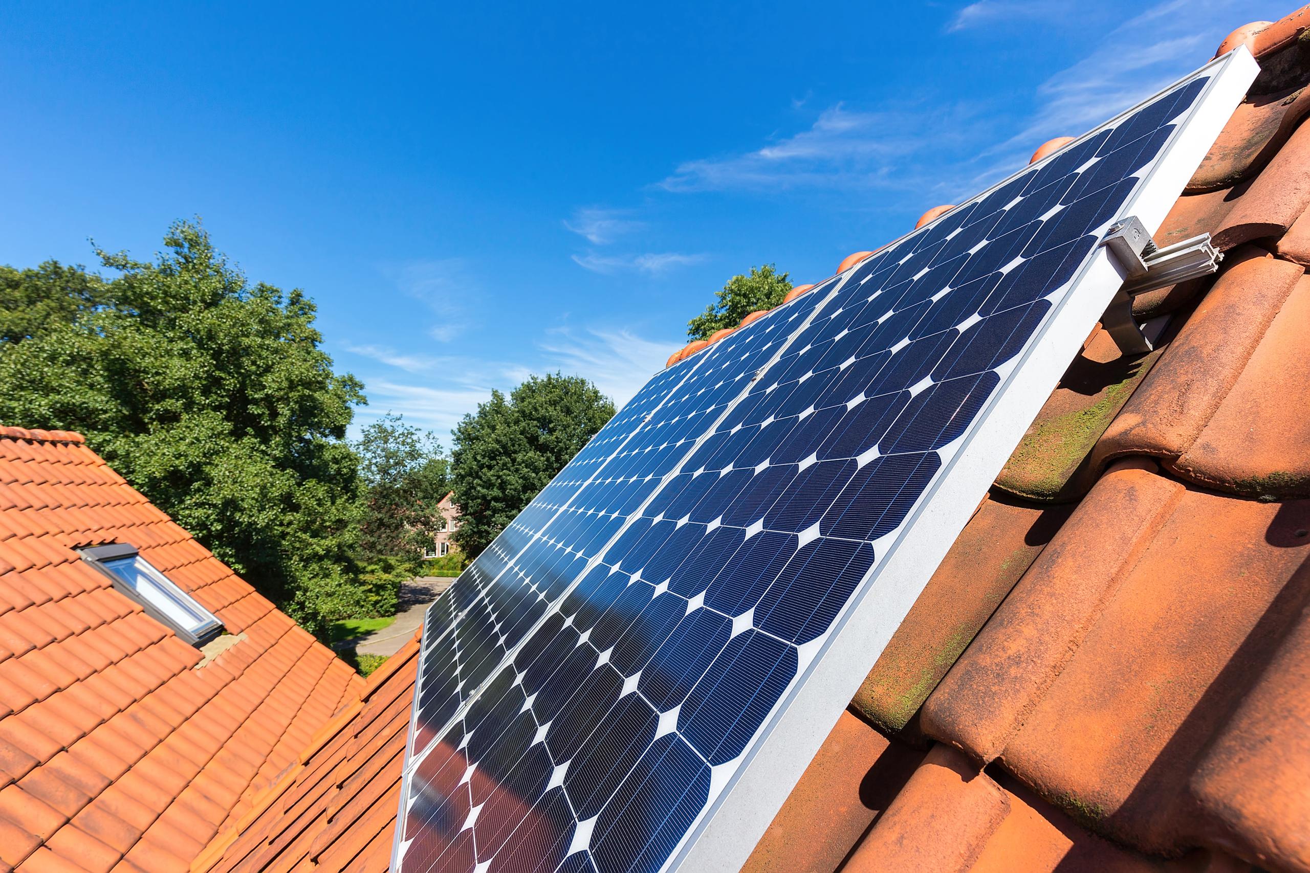 A row of blue solar panels on a tiled house roof with trees and blue sky in the background