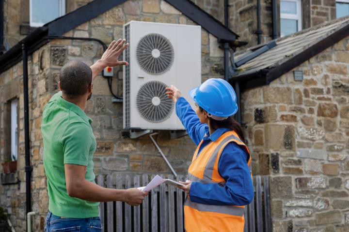 installation worker holding a digital tablet and househoder pointing to an air source heat pump being installed on the side of a home
