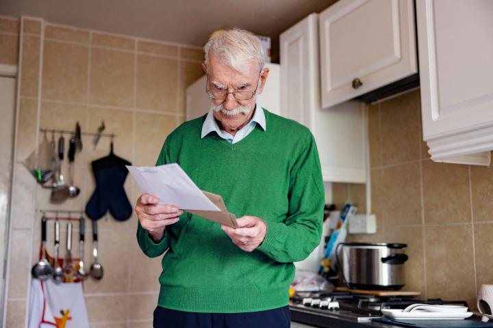 elderly man standing in his kitchen taking an energy bill out of a brown paper envelope