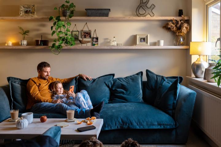 a father relaxing on the sofa with his daughter leaning against him, holding a mobile phone
