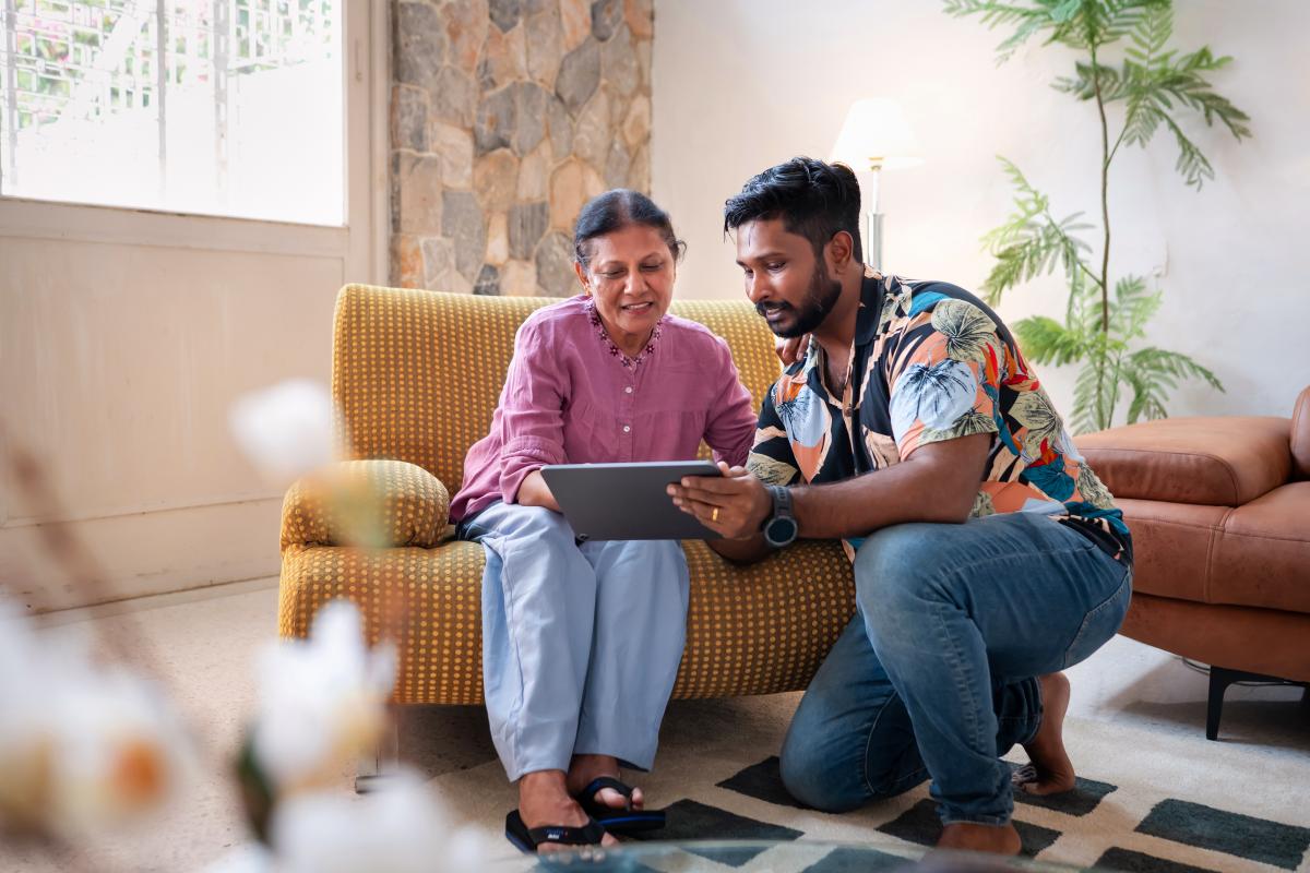 a mid-aged South Asian man holding a digital tablet for his mother who is sitting on a colourful sofa